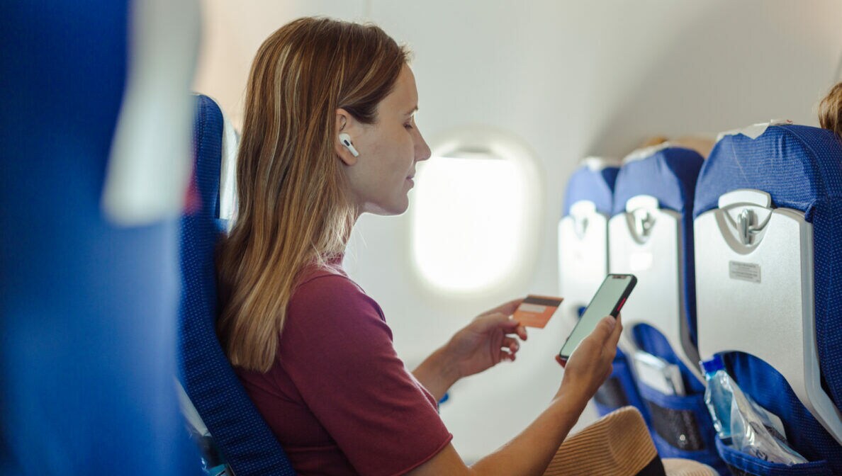 Eine Frau sitzt in einem Flugzeug mit dem Smartphone in der einen Hand und einer Geldkarte in der anderen Hand.