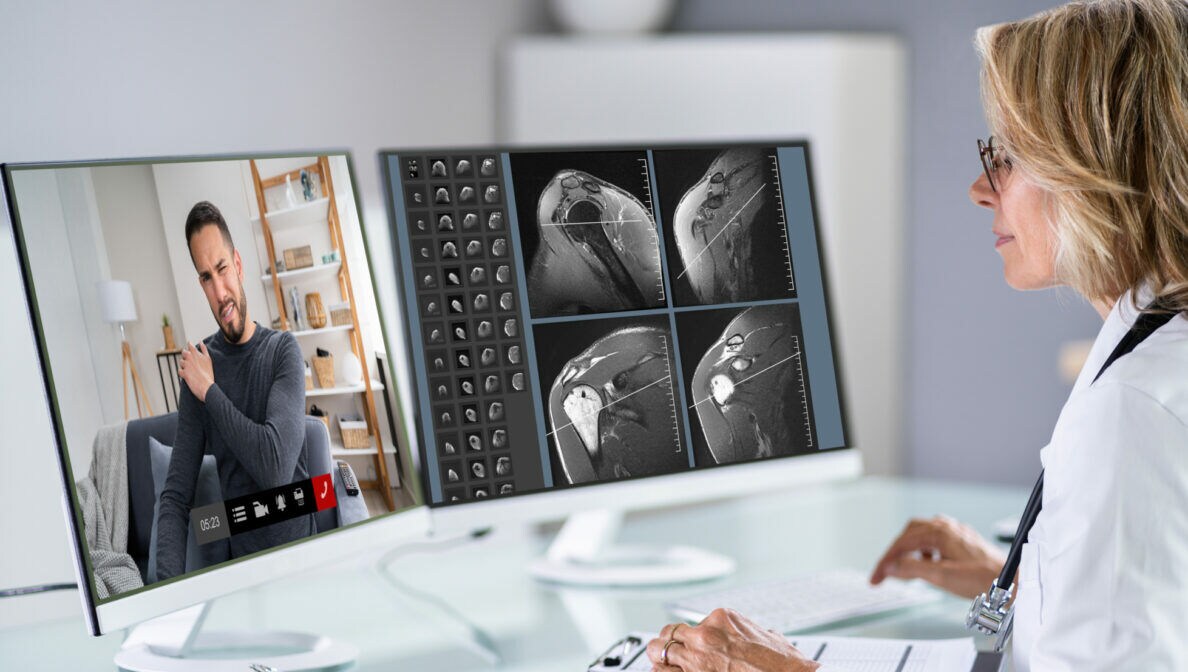 Eine Ärztin sitzt an ihrem Schreibtisch vor zwei Bildschirmen und kommuniziert mit einem Patienten via Videotelefonie.