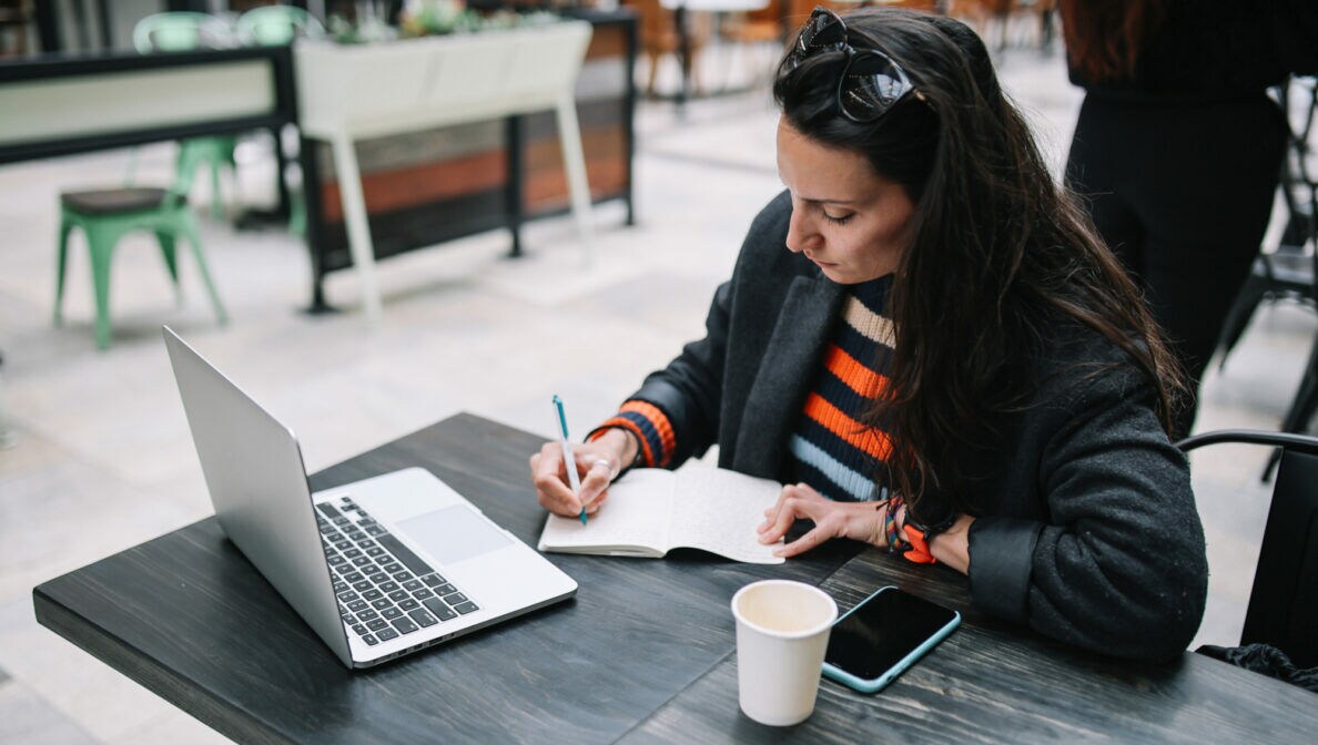 Eine Frau sitzt an einem Tisch vor einem aufgeklappten Laptop und schreibt mit einem Stift in ein geöffnetes Notizbuch.