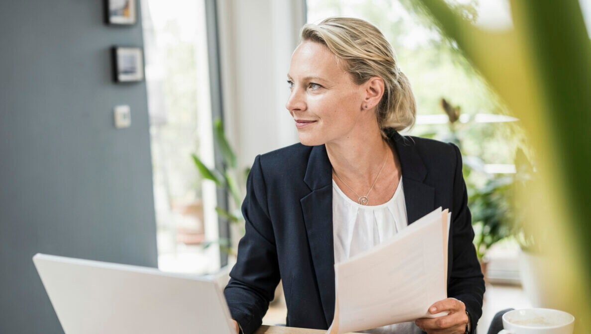 Eine Frau sitzt lächelnd an einem Schreibtisch vor einem Laptop und hält in der linken Hand ein Bündel von Papieren.
