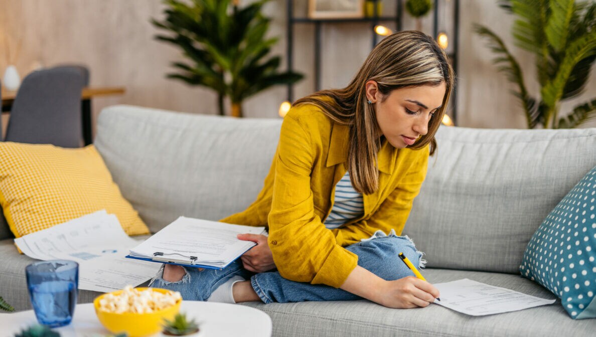 Eine Frau mit gelber Hemdjacke sitzt auf einem Sofa und füllt Dokumente aus.
