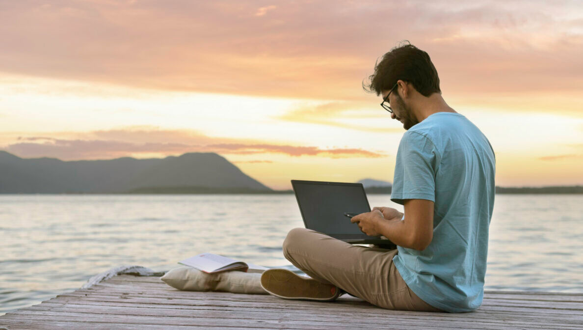Eine Person sitzt bei Sonnenuntergang mit einem Laptop und einem Buch am Steg und tippt