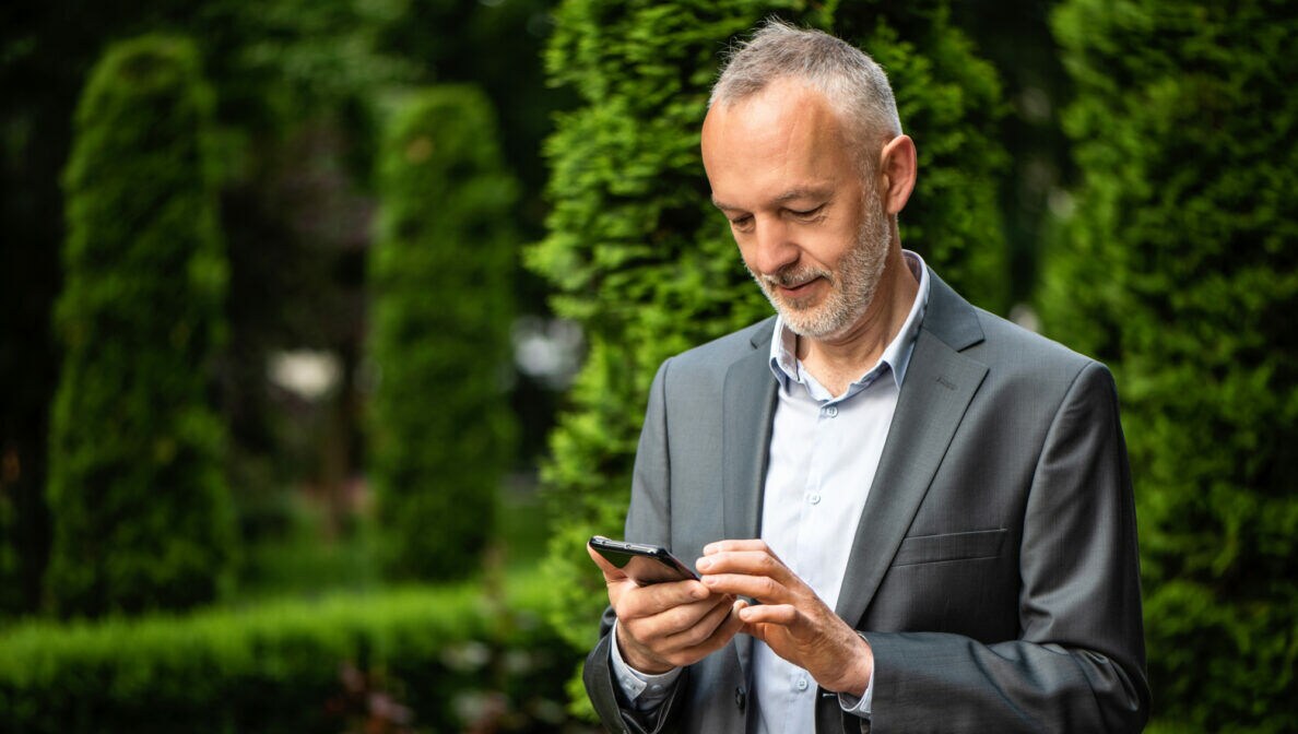 Ein Mann steht vor einer Hecke und schaut auf sein Smartphone.