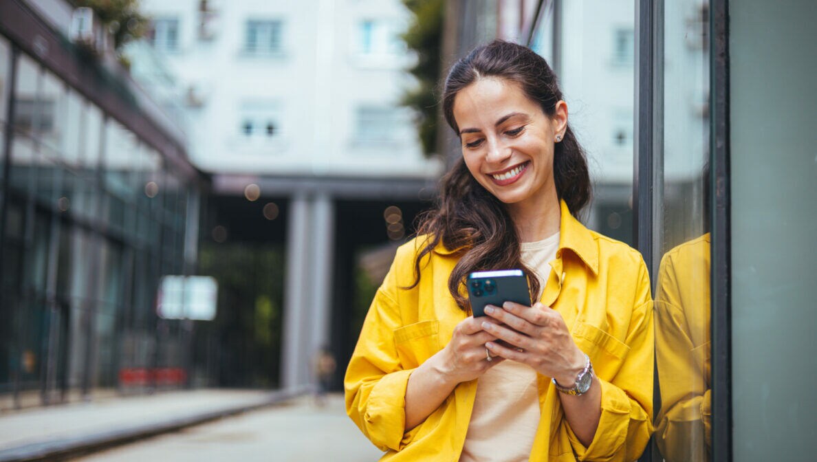 Eine Person lehnt sich mit der linken Schulter an eine Glasfassade und schaut lächelnd auf ihr Smartphone, das sie in den Händen hält.