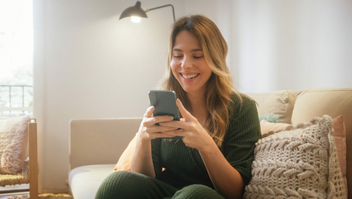 Eine Person sitzt auf einem Sofa und schaut lachend auf das Smartphone, das sie mit beiden Händen festhält.