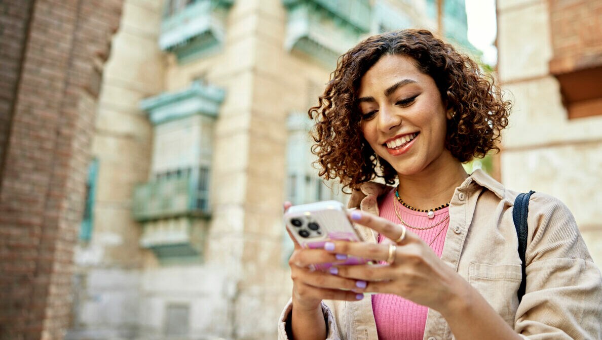 Eine Person steht zwischen hohen Steingebäuden und blickt auf das Smartphone in ihren Händen.