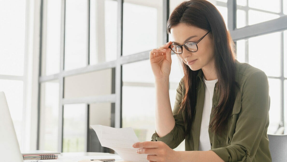 Eine junge Frau sitzt am Schreibtisch mit einem Laptop und Taschenrechner in einem hellen Büro. Dabei schaut sie durch ihre Brille auf ein Dokument, das sie in der Hand hält.