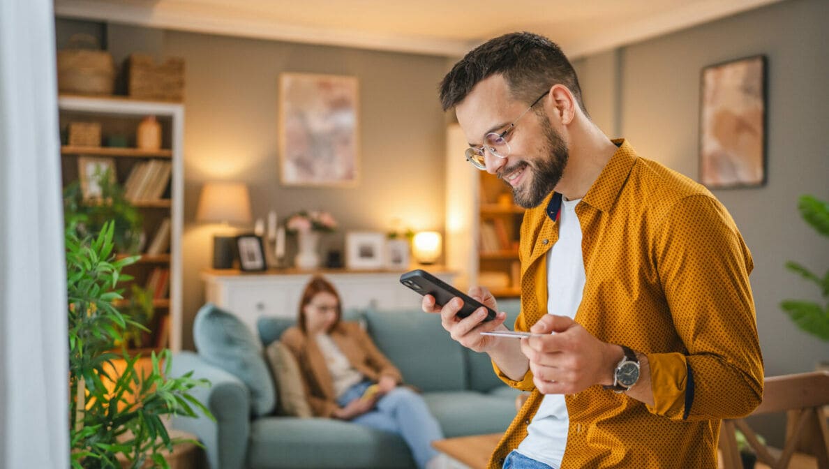 Eine Person sitzt auf einem Tisch und hält eine Kreditkarte sowie ein Smartphone in den Händen. Im Hintergrund sitzt eine weitere Person auf einem Sofa.