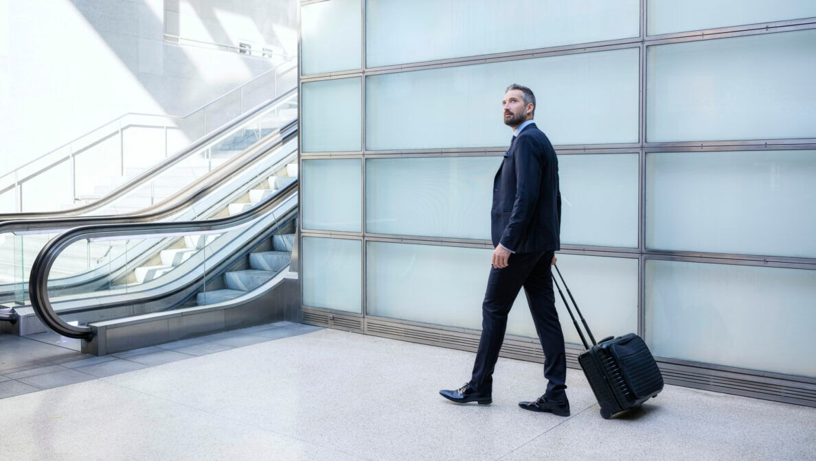 Ein Mann im Anzug und mit einem Rollkoffer steht in der Nähe einer Rolltreppe.