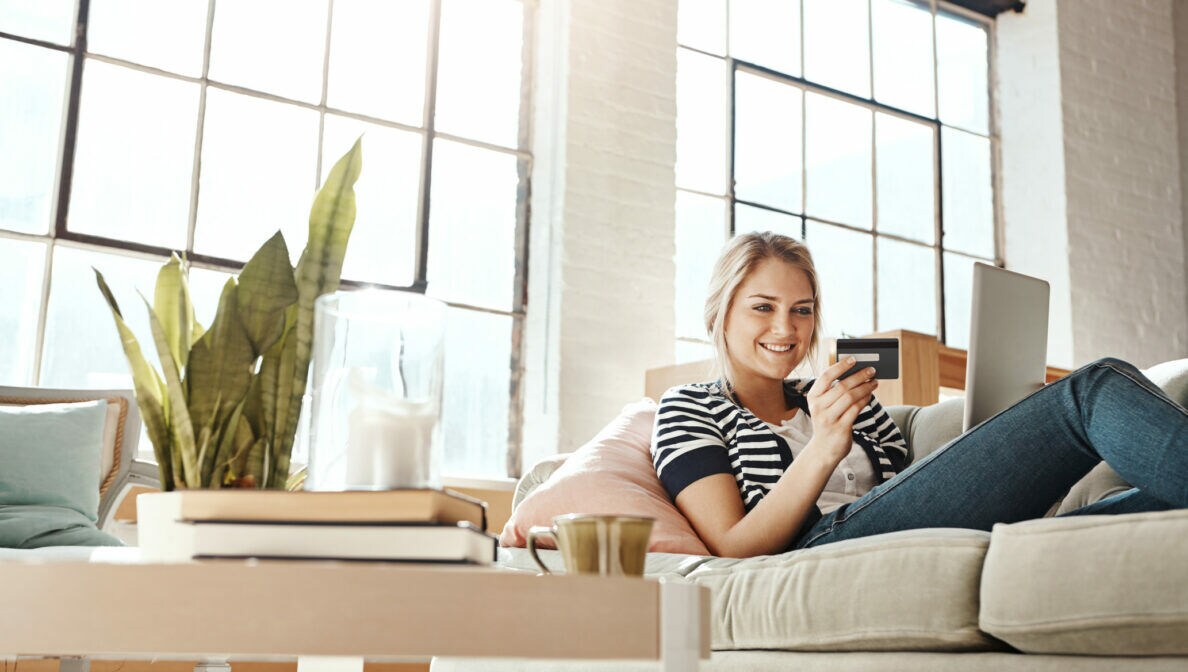 Eine Person liegt auf einem Sofa, hat ein Laptop auf ihrem Schoß und blickt lächelnd auf eine Bankkarte.