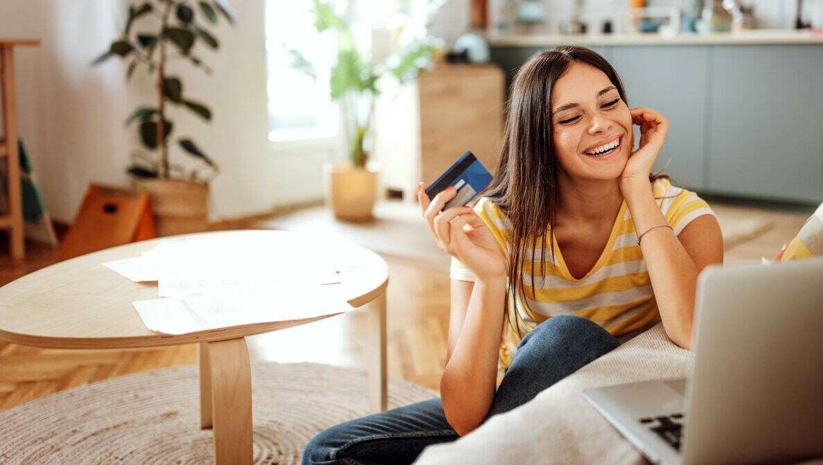 Eine Person sitzt auf dem Fußboden zwischen Couchtisch und Sofa, hält eine Kreditkarte in der Hand und blickt auf ein geöffnetes Notebook, das auf der Sofasitzfläche steht. 