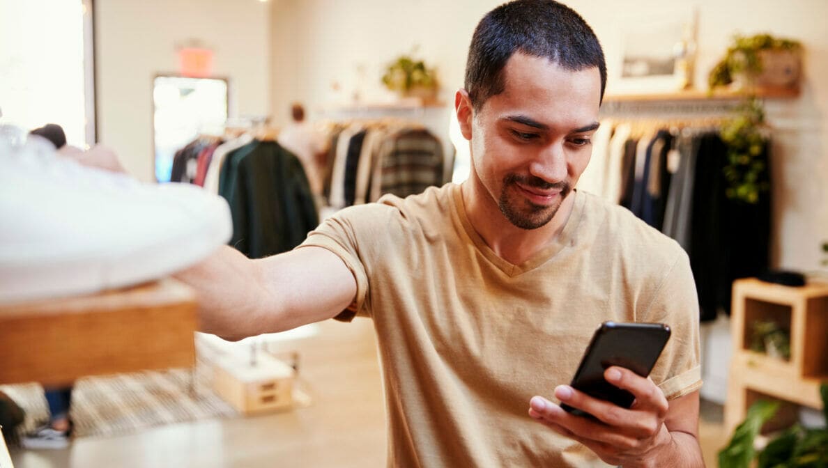 Eine Person steht in einem Bekleidungsgeschäft, greift mit einer Hand nach einem Paar Sneaker und hält in der anderen Hand ein Smartphone.