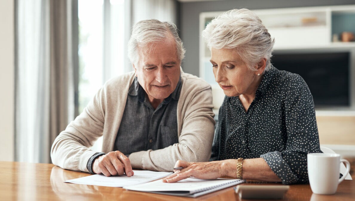 Eine ältere Frau und ein älterer Mann sitzen am Tisch. Sie schauen sich aufmerksam Dokumente an, die vor ihnen liegen.