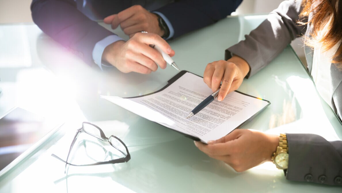 Zwei Personen an einem Tisch zeigen mit Finger und Stift auf ein Dokument in einem Klemmbrett.