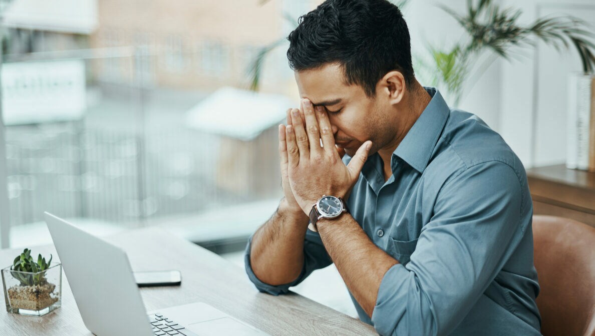 Ein Mann sitzt an einem Schreibtisch vor einem aufgeklappten Laptop mit geschlossenen Augen, stützt die Ellenbogen auf den Tisch und drückt die Zeigefinger an die Nasenwurzel.