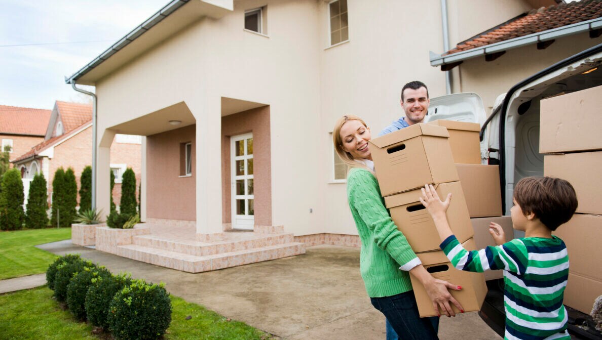 Ein Mann, eine Frau und ein Kind stehen Kisten tragend an einem offenen Transporter vor einem Haus.