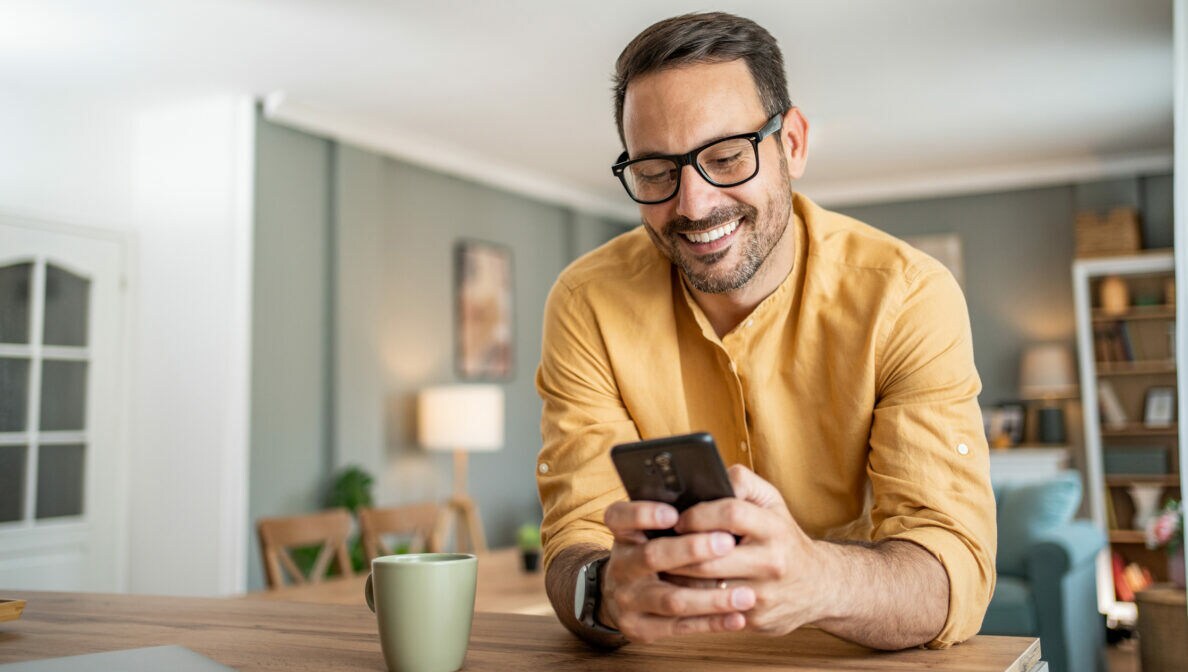 Eine Person lehnt auf einer Kücheninsel und blickt lächelnd auf das Smartphone in ihren Händen.