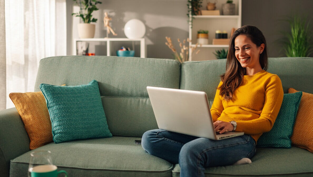 Eine Person sitzt auf einem Sofa und schaut auf das geöffnete Notebook vor sich.