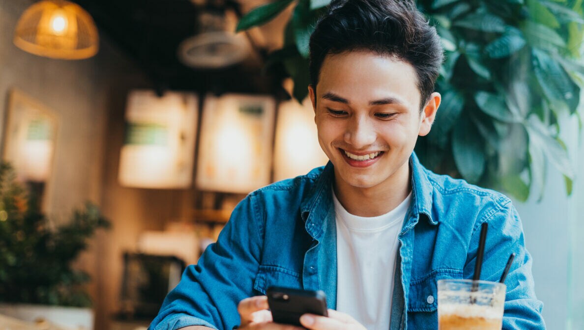 Eine Person sitzt mit einem Café vor sich am Tisch und blickt auf das Smartphone, das sie in den Händen hält.