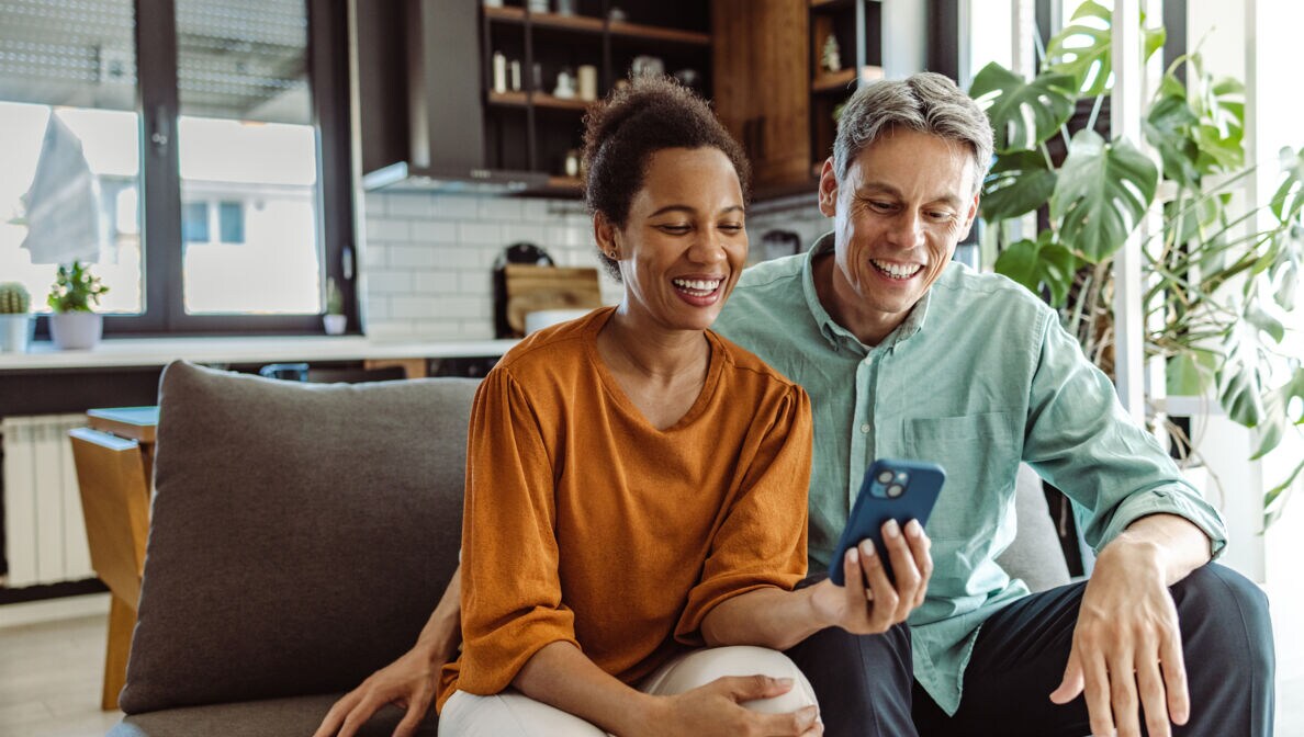 Zwei Personen sitzen gemeinsam auf einem Sofa und blicken zusammen auf ein Handy.