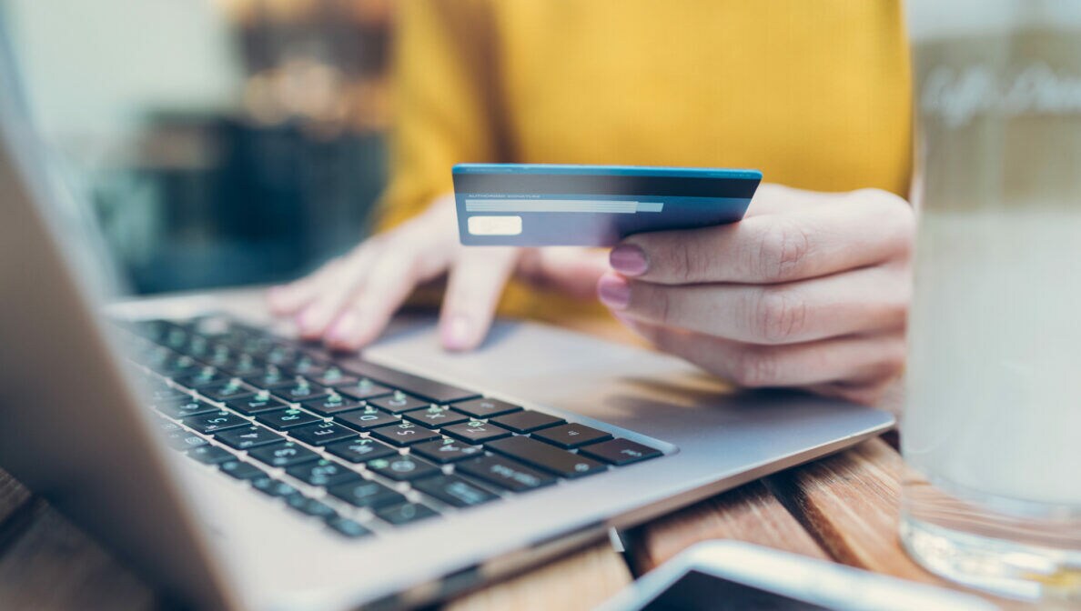 Nahaufnahme einer Person vor einem geöffneten Laptop, in einer Hand eine Bankkarte haltend, die andere liegt auf der Laptoptastatur.