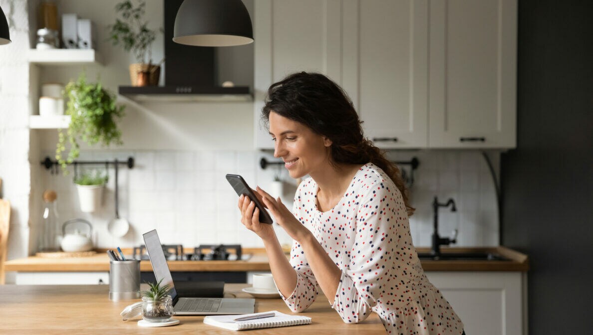 Eine Person lehnt auf einer Kücheninsel und blickt auf das Smartphone in ihren Händen. Vor ihr steht ein geöffnetes Laptop.