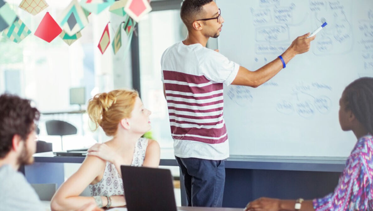 Mann steht mit einem Stift in der Hand an einem Whiteboard und zeigt seinen drei Arbeitskolleg:innen seine Notizen.