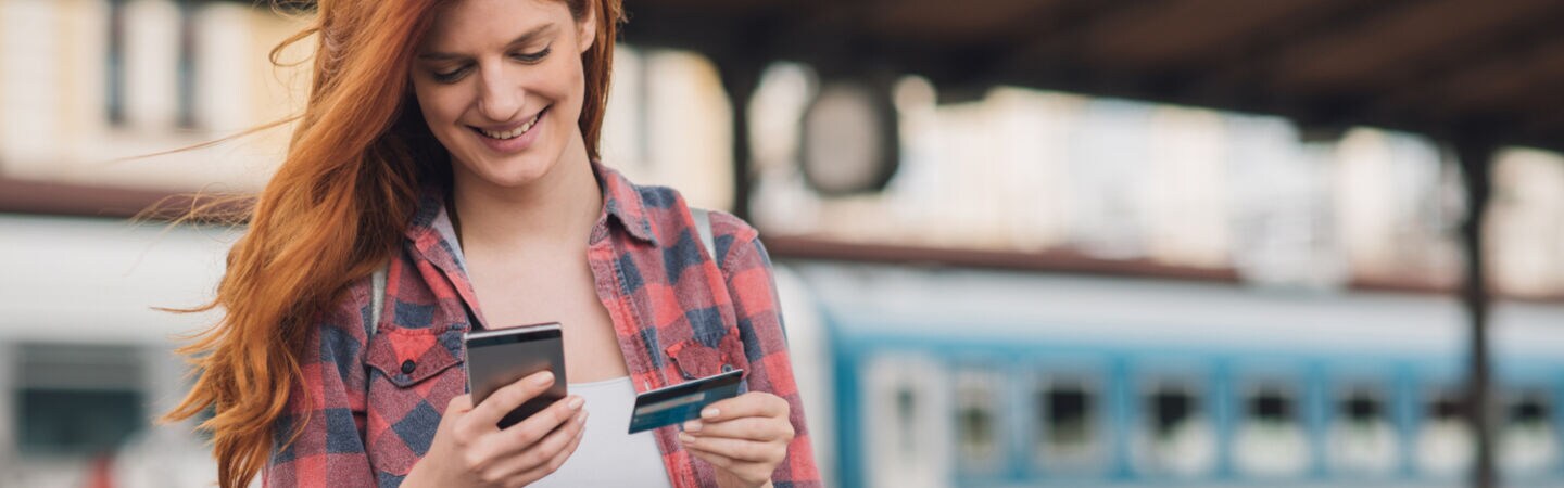 Eine lächelnde junge Frau am Bahngleis mit Smartphone in der einen, Kreditkarte in der anderen Hand