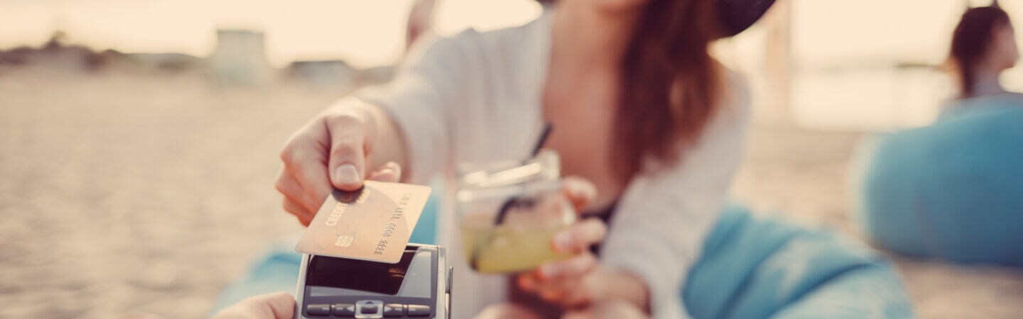 Eine Frau mit Sonnenbrille, Hut und Drink in der Hand am Strand bezahlt per Kreditkarte an einem mobilen Zahlterminal