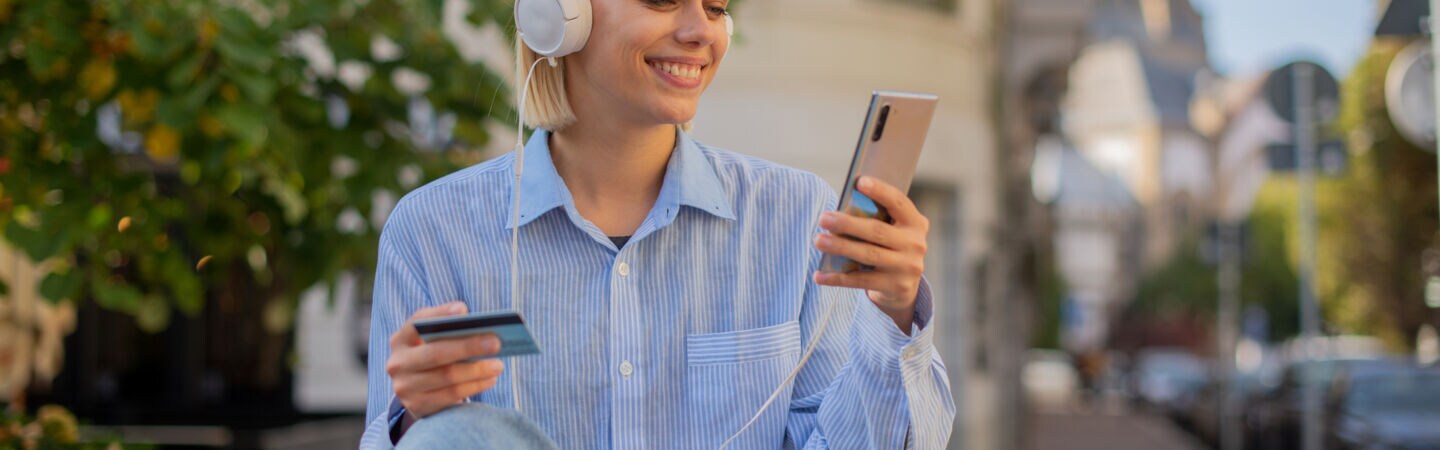 Eine Person sitzt im Freien auf einer Bank, hat Kopfhörer auf und hält ein Smartphone sowie eine Bankkarte in ihren Händen.