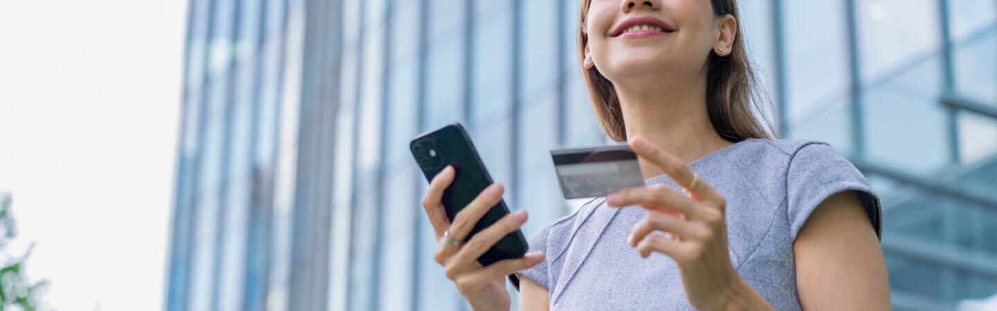 Eine lächelnde Frau steht mit Handy und Kreditkarte in der Hand vor einem Gebäude