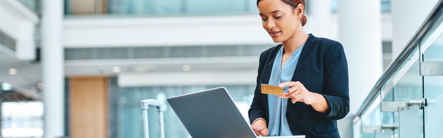 Eine Person sitzt in einem Bürogebäude, hat einen Koffer neben sich stehen und ein Laptop auf den Knien. In ihrer Hand hält sie eine Bankkarte.