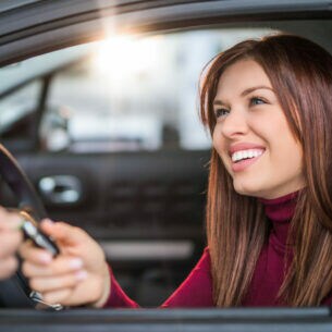 Eine lächelnde Frau im Auto bekommt von einer anderen Person durchs Fenster einen Autoschlüssel gereicht