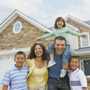 Eine Frau, ein Mann und drei Kinder, eins davon auf den Schultern des Mannes, lachen in die Kamera. Im Hintergrund steht ein Einfamilienhaus.