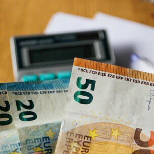 Nahaufnahme mehrerer Geldscheine, im Hintergrund Taschenrechner, Stift und Papier auf einem Schreibtisch