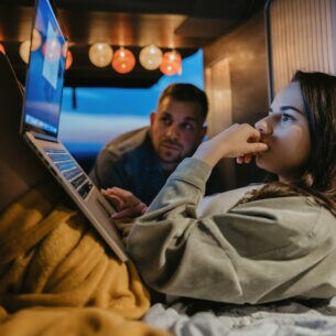 Junge Frau bei Dämmerung in einem offenen Van mit Laptop auf den Beinen und ein Mann, der ebenfalls auf den Bildschirm blickt