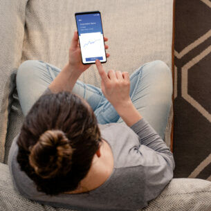 Junge Frau sitzt auf einem Sofa und schaut auf eine Trading-App auf ihrem Smartphone.