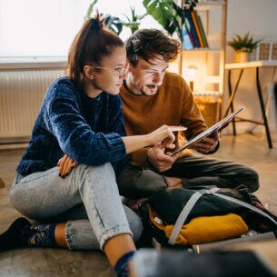 Eine junge Frau und ein junger Mann, in einer Wohnung auf dem Fußboden sitzend, die auf ein Tablet schauen, das er in den Händen hält
