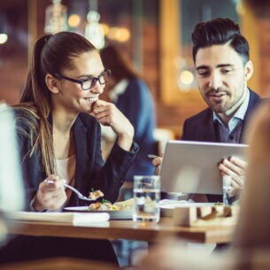 Zwei Personen in Anzügen sitzen gemeinsam in einem Restaurant und unterhalten sich, während sie auf ein Tablet blicken.