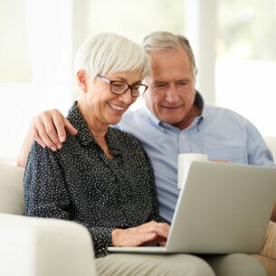 Eine ältere Frau und ein älterer Mann gemeinsam auf dem Sofa und auf ein Laptop blickend, der auf ihren Knien steht