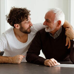 Ein lachender Mann mit grauem Bart und grauen Haaren wird von einem lachenden anderen Mann umarmt.