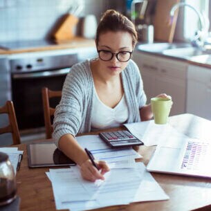 Eine Frau mit Brille und einer Kaffeetasse in der Hand sitzt an einem Küchentisch und bearbeitet Dokumente.