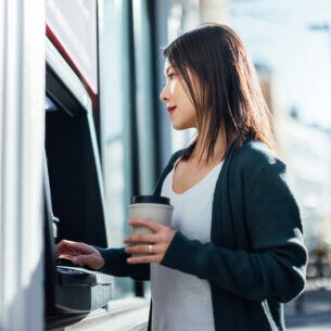 Eine junge Frau mit einem Becher in der einen Hand in einer Straße vor einem Geldautomaten