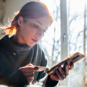 Eine junge Frau mit Zopf und Rollkragenpullover sitzt zu Hause und schaut auf das Smartphone in ihrer linken Hand. In ihrer rechten Hand hält sie eine Bankkarte.