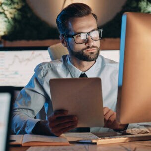 Ein Mann mit Brille am Schreibtisch, der ein Tablet in der Hand hält und auf einen Computerbildschirm blickt