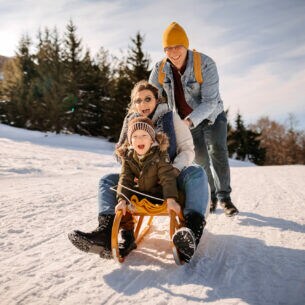 Eine dreiköpfige Familie im Schnee, von der zwei Personen auf einem Schlitten sitzen und die dritte Person anschiebt.