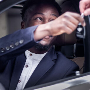 Eine Person sitzt auf dem Fahrersitz eines Autos, und nimmt durch das offene Fenster von einer anderen Person einen Autoschlüssel entgegen
