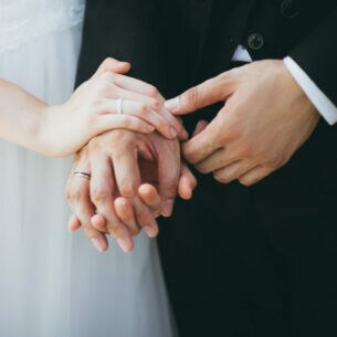 Nahaufnahme von Händen mit Eheringen. Sie gehören einer Frau im Brautkleid sowie einem Mann im Anzug.