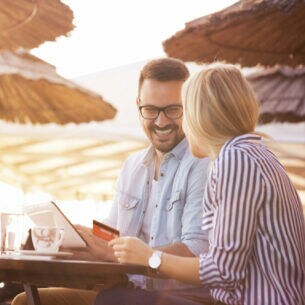 Junges Paar sitzt lachend unter Strandschirmen mit Tablet und Kreditkarte.