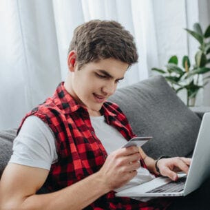 Eine junge Person sitzt vor einem aufgeklappten Laptop auf dem Sofa und hält eine Kreditkarte in der Hand.